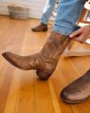 A person pulling on a brown cowboy boot while standing on a wooden floor, with another boot nearby.