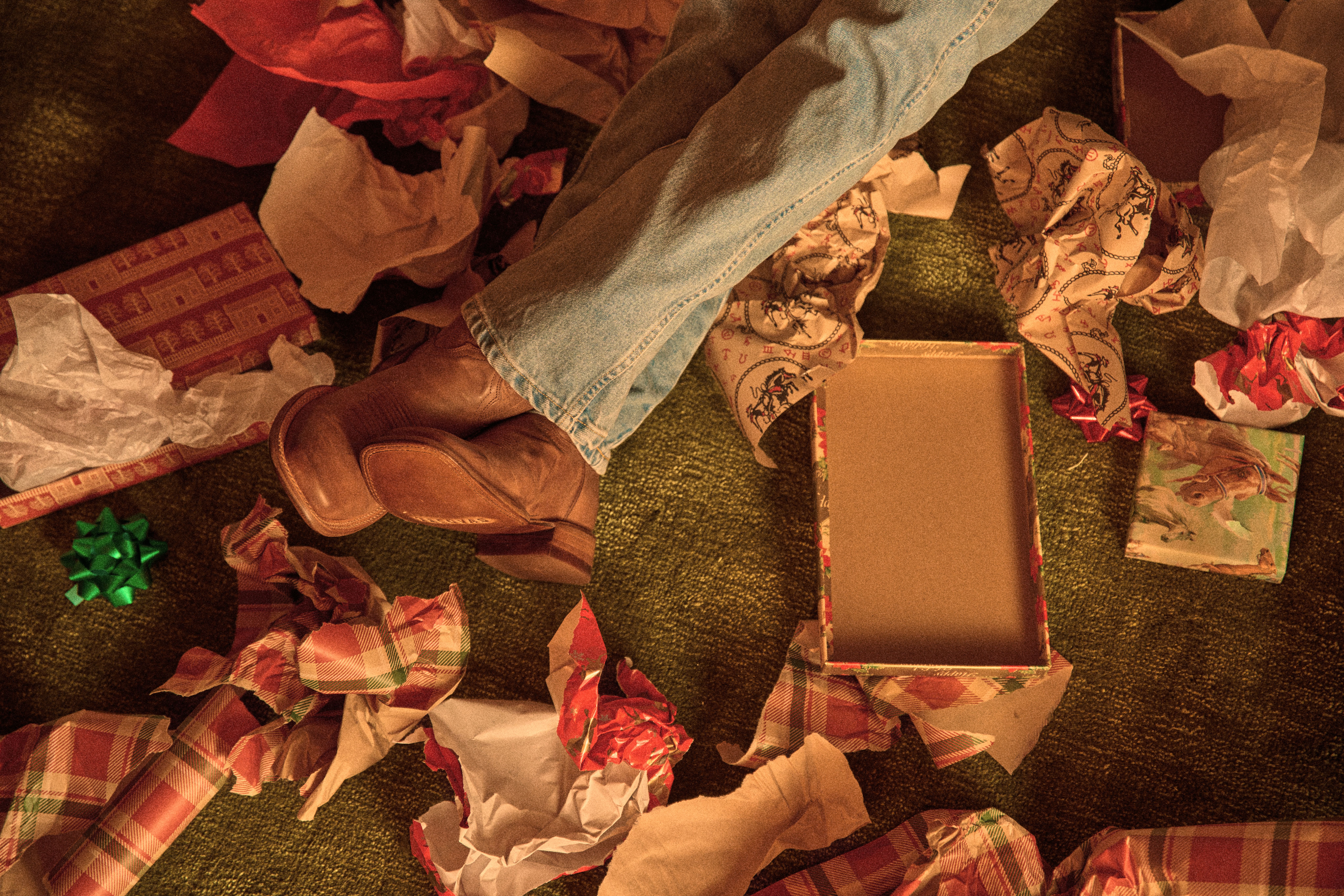 Person wearing jeans and brown boots laying in a pile of torn gift wrap, an open gift box, unwrapped presents, and a green bow on a textured green carpet.