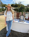 A woman wearing a hat and white blouse stands beside a table displaying cowboy boots outdoors on a sunny day.