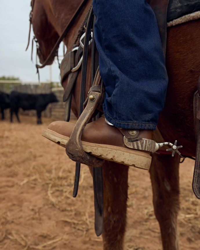 A person wearing jeans and brown boots with spurs is sitting on a horse. Cattle are seen in the background on a dirt surface.