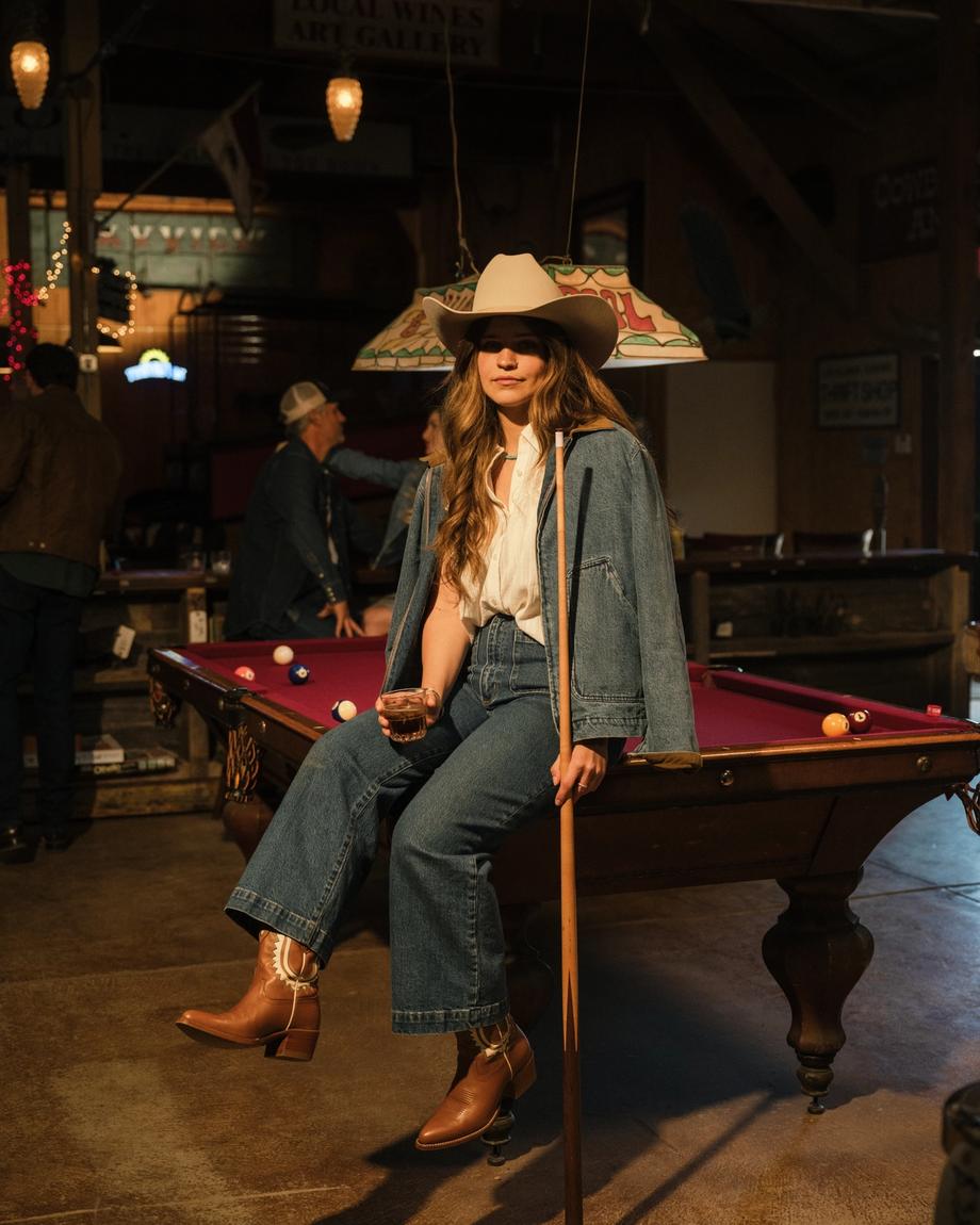 A person in a cowboy hat and denim outfit sits on a pool table holding a cue stick in a dimly lit room.