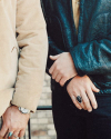 Two people standing side by side, both wearing jackets. One wears a beige jacket and the other a dark leather jacket. Each person has large rings on their fingers and one person wears a wristwatch.