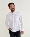 A bearded man stands against a plain background wearing a white button-down shirt and jeans, with one hand in his pocket.