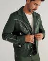 A man wearing a dark green embroidered jacket over a white shirt fastens the buttons while standing against a plain light background.