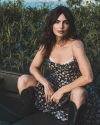 A woman in a floral dress and black boots sits outdoors near tall grass, with water in the background.