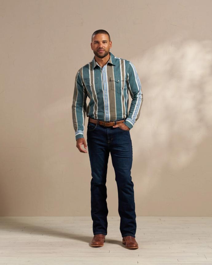 A man stands against a beige wall wearing a striped button-up shirt, dark blue jeans, and brown boots. His right hand rests slightly in his pocket.