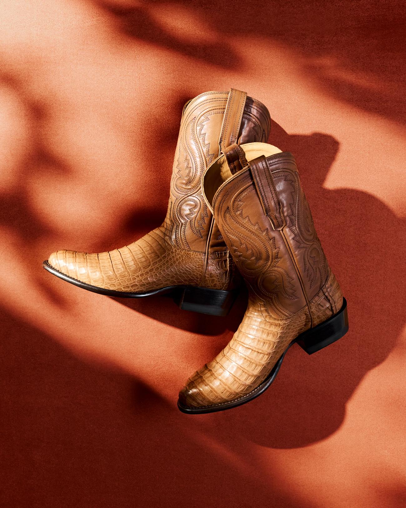 Close-up of a person's feet wearing brown crocodile-patterned cowboy boots. The boots are resting on the step of a green vehicle, with grass visible in the background.