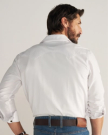 A man with dark hair and a beard is shown from the back, wearing a white collared shirt and blue jeans with a brown leather belt.