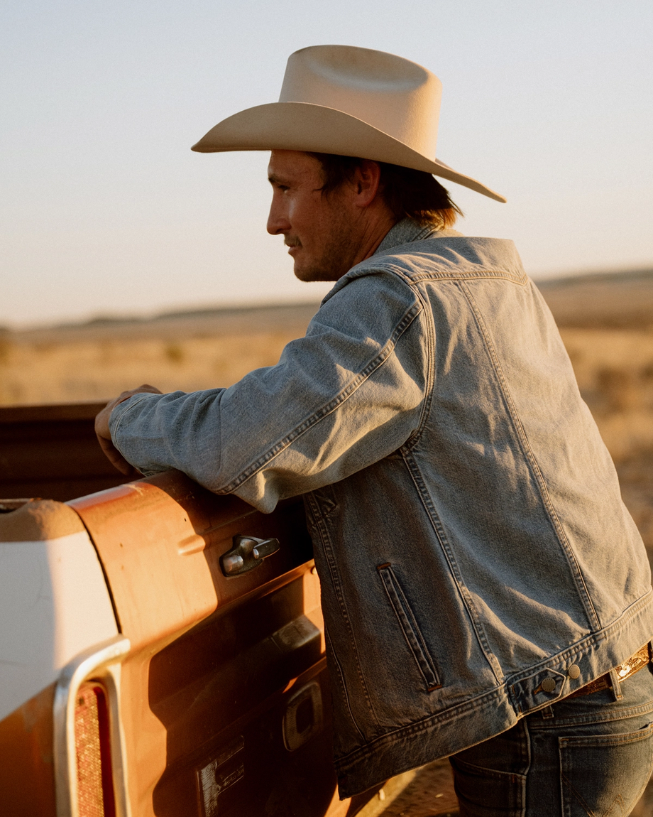 A man in a denim jacket and cowboy hat leans on the back of a dusty pickup truck in a dry, open landscape.