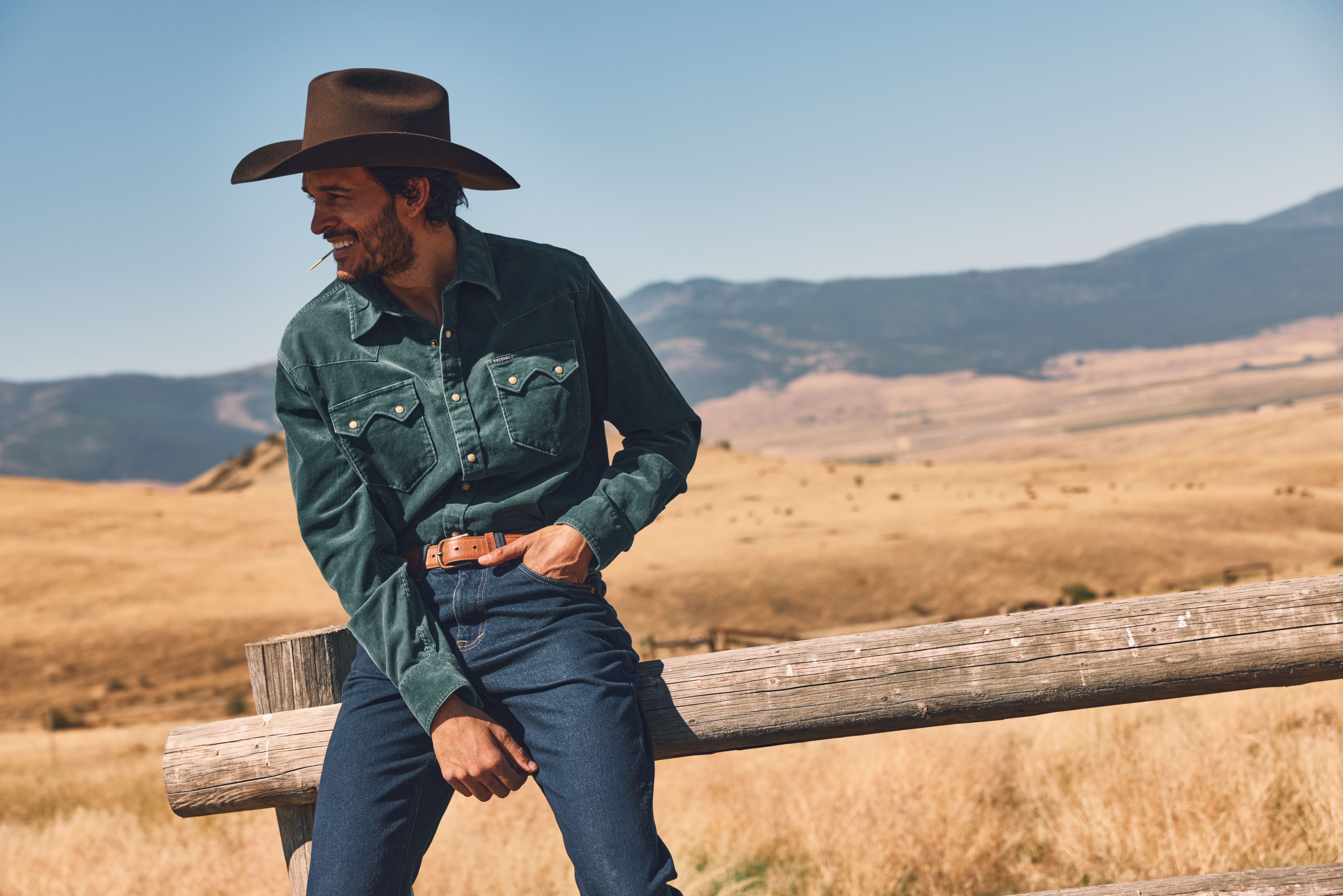 Man in a cowboy hat and denim outfit leans on a wooden fence in a dry, open landscape with hills in the background.