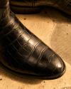 Close-up of a dark leather shoe with a crocodile skin texture on a patterned stone floor.