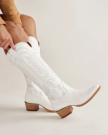 A person pulls on a pair of tall white cowboy boots with wooden heels and decorative stitching, sitting against a plain light background.