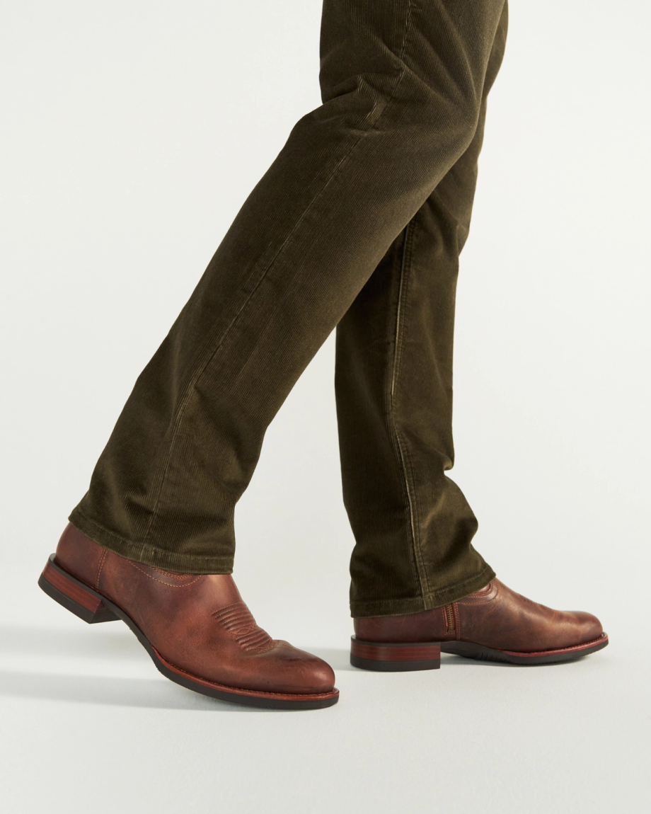 The Wayne in Maplewood Cowhide. A person wearing The Wayne Cowhide brown leather shoes and olive green pants is shown mid-step against a plain background.