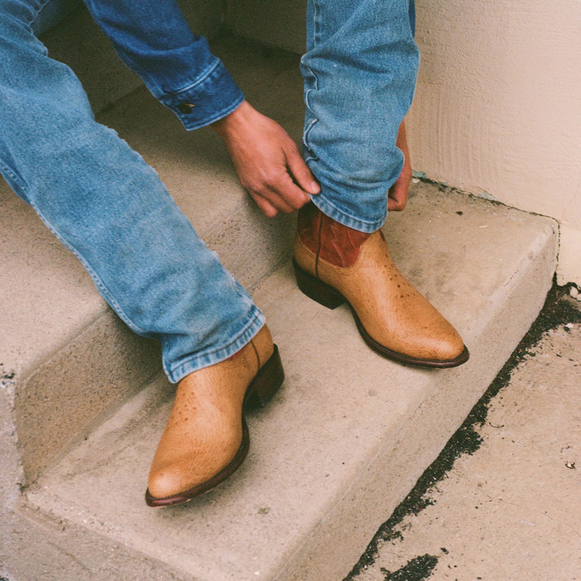Person sitting on concrete steps adjusts the hem of blue jeans, wearing tan cowboy boots with brown accents.
