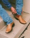 Person sitting on concrete steps adjusts the hem of blue jeans, wearing tan cowboy boots with brown accents.