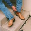 Person sitting on concrete steps adjusts the hem of blue jeans, wearing tan cowboy boots with brown accents.