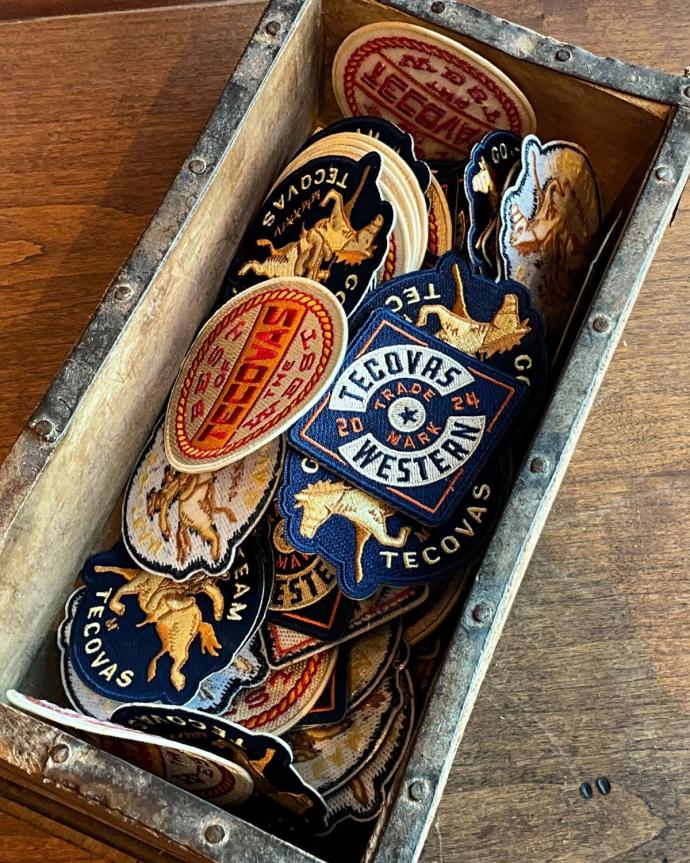 A wooden box filled with assorted embroidered patches, mostly with "Tecovas" and western-themed designs, placed on a wooden surface.