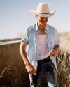 Man wearing a cowboy hat, light blue shirt, and jeans stands outdoors in dry grass under a clear sky, with his shirt wet and a slight smile on his face.