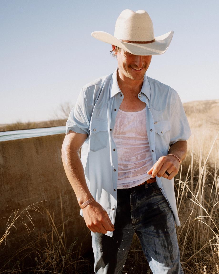 Man wearing a cowboy hat, light blue shirt, and jeans stands outdoors in dry grass under a clear sky, with his shirt wet and a slight smile on his face.