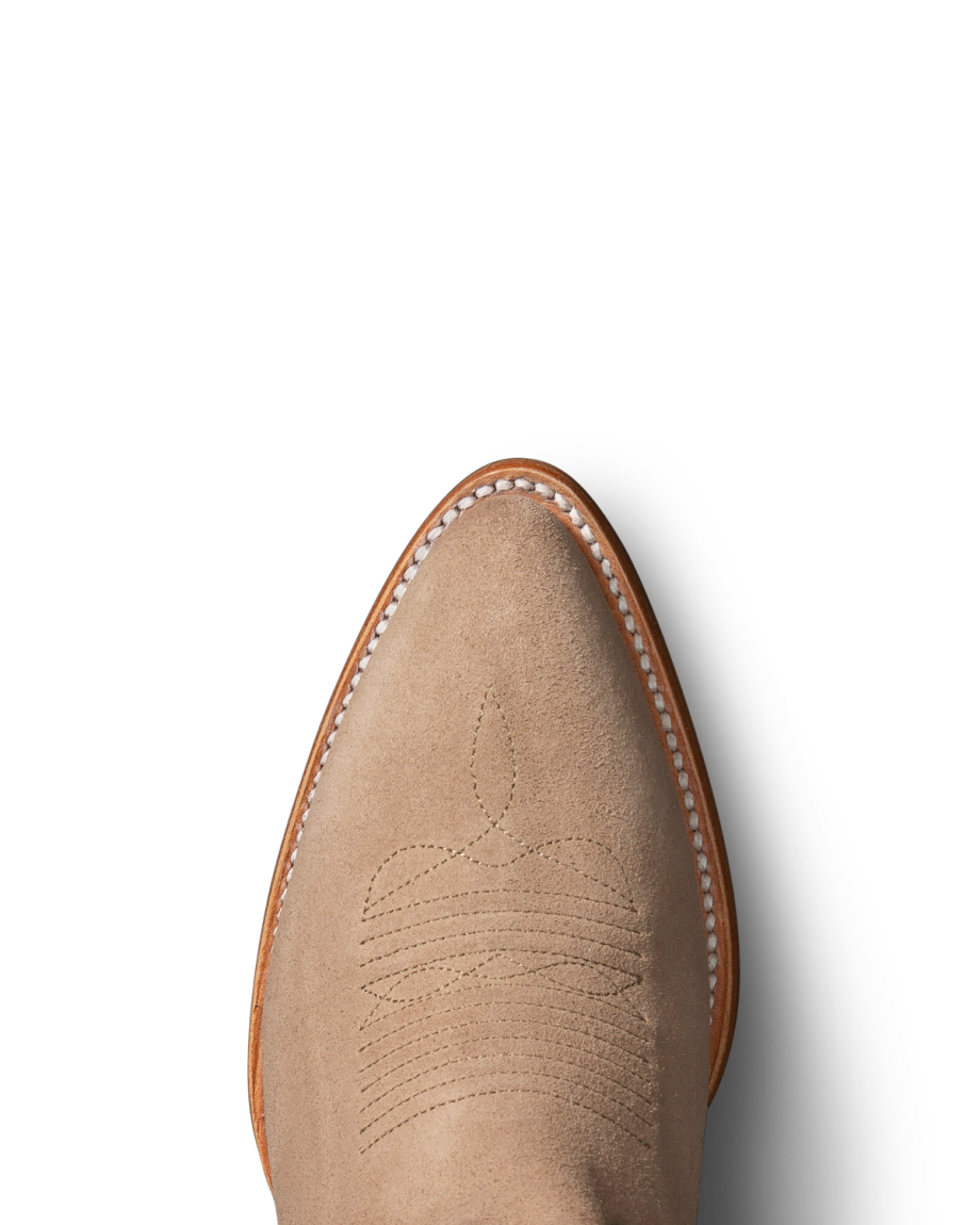 Close-up of a tan suede cowboy boot on a black background, featuring detailed stitching on the upper part and white trim along the edge.