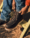 Person lacing up brown leather work boots while sitting on a yellow chair outdoors, wearing blue jeans and a red shirt.