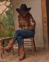 A person wearing a cowboy hat, brown sweater, jeans, and cowboy boots sits on a wooden chair in a rustic outdoor setting.