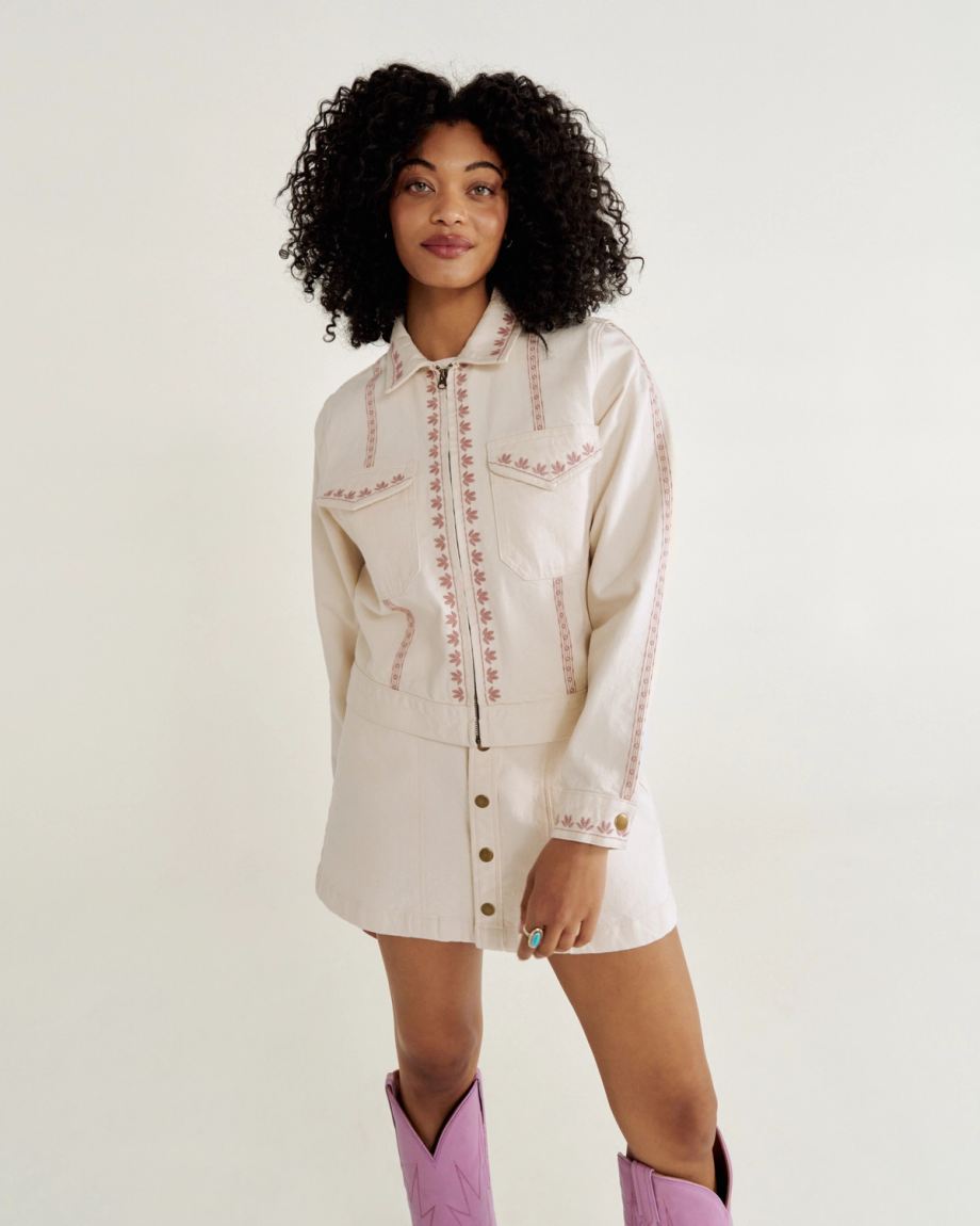Person with curly hair wearing a light-colored jacket and skirt set with embroidery and pink cowboy boots, standing against a plain background Natural Denim .