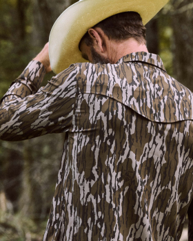 A man in a camouflage shirt and cowboy hat stands outdoors, facing away and adjusting his hat, with trees in the background.