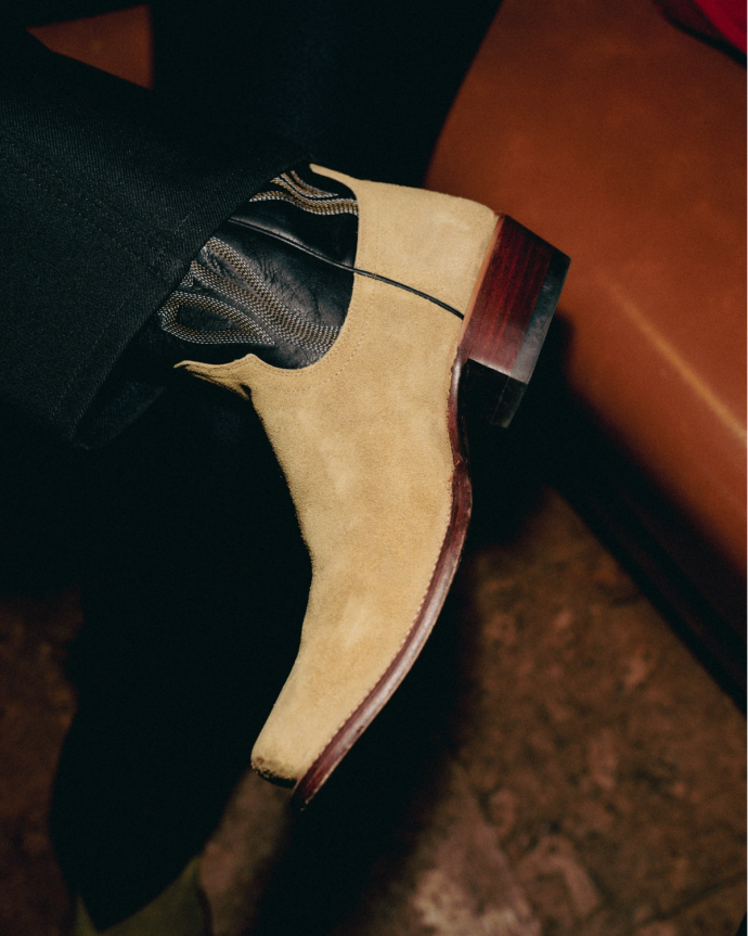 Beige suede ankle boot with a brown wooden heel, worn with black trousers and socks with a zebra pattern.