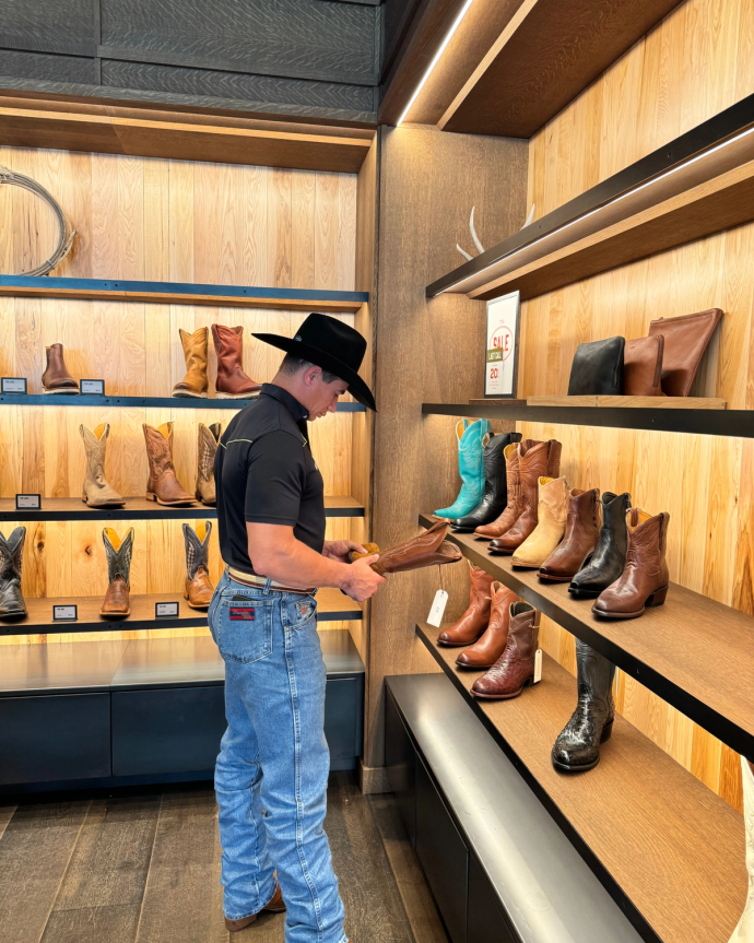 A man in a black hat and jeans examines a brown boot in a store. Several other boots are displayed on wooden shelves around him.