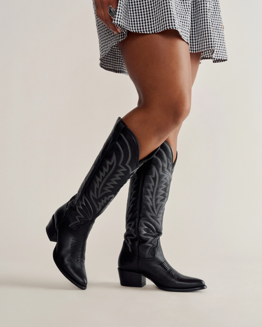 A person wearing a black and white checkered skirt and black knee-high cowboy boots stands against a plain background.