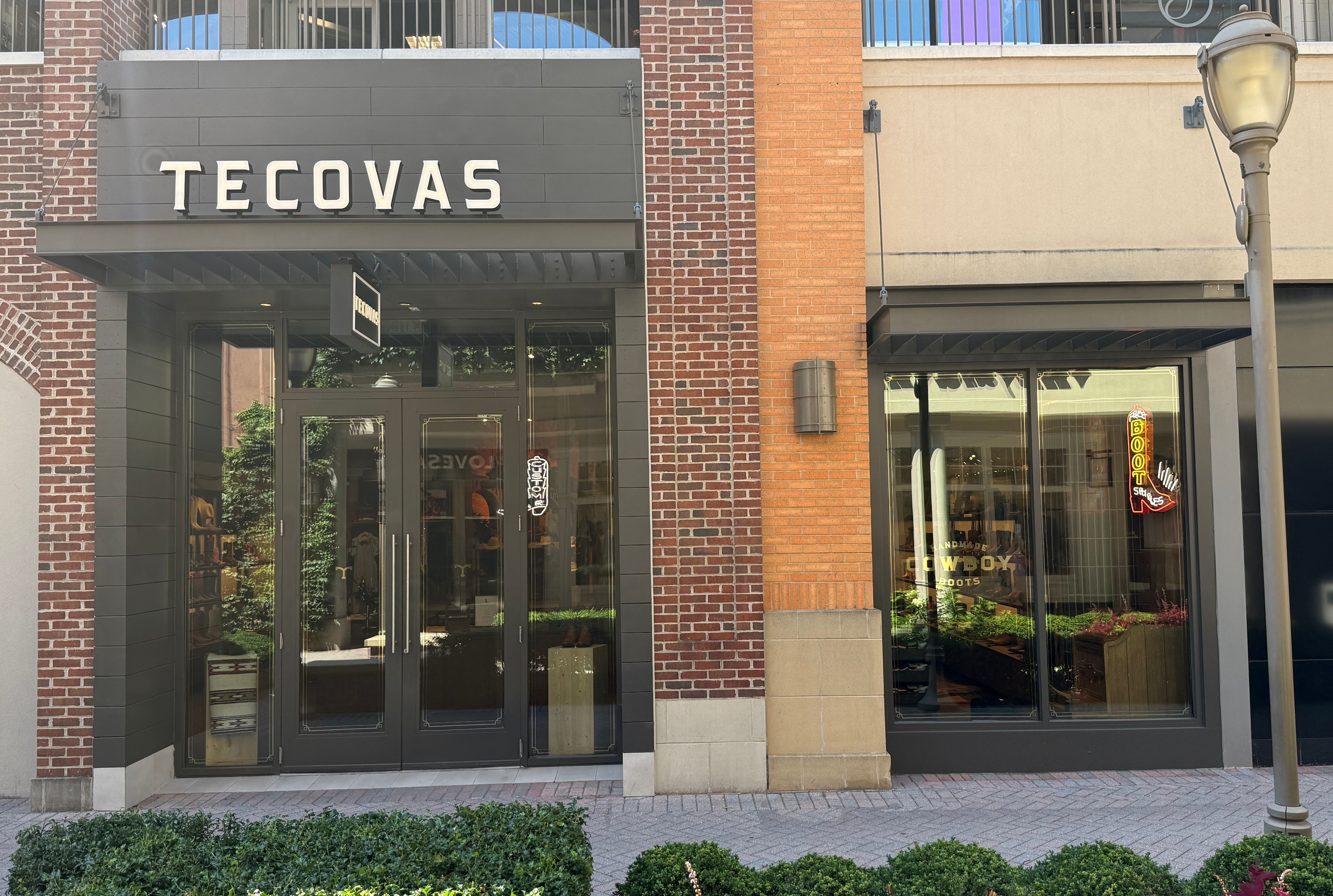 A storefront with a “Tecovas” sign above glass double doors, next to large display windows showing boots inside. Brick and beige building exterior, with a streetlamp and shrubs in front.