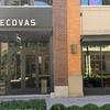 A storefront with a “Tecovas” sign above glass double doors, next to large display windows showing boots inside. Brick and beige building exterior, with a streetlamp and shrubs in front.