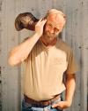 A bearded man in a tan t-shirt and jeans holds a camouflage cap to his head, squinting one eye, standing against a corrugated metal wall.