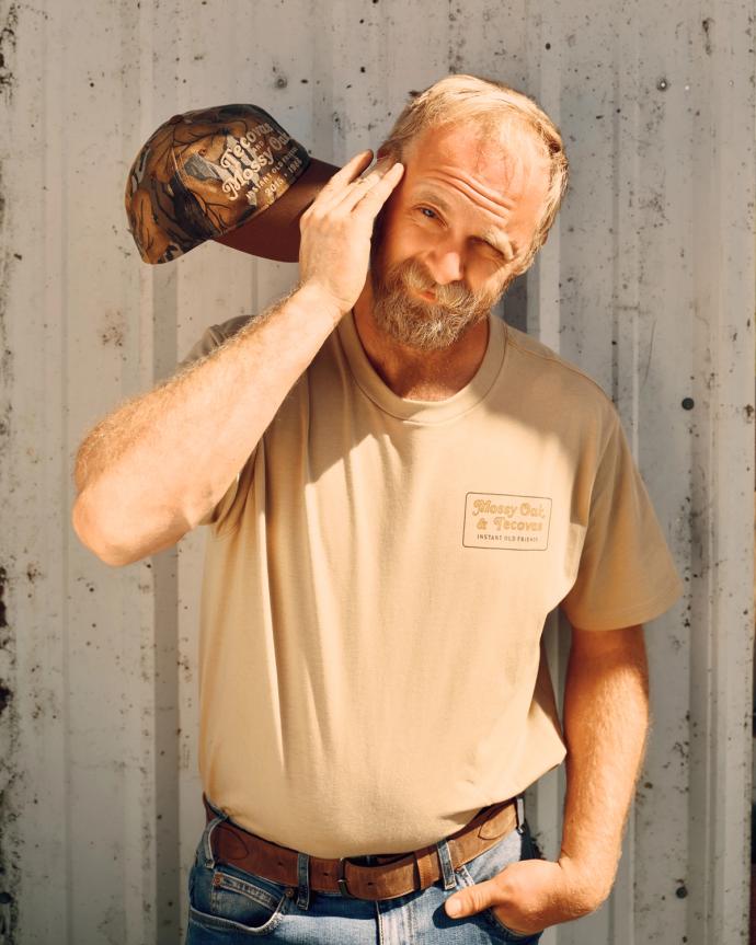 A bearded man in a tan t-shirt and jeans holds a camouflage cap to his head, squinting one eye, standing against a corrugated metal wall.