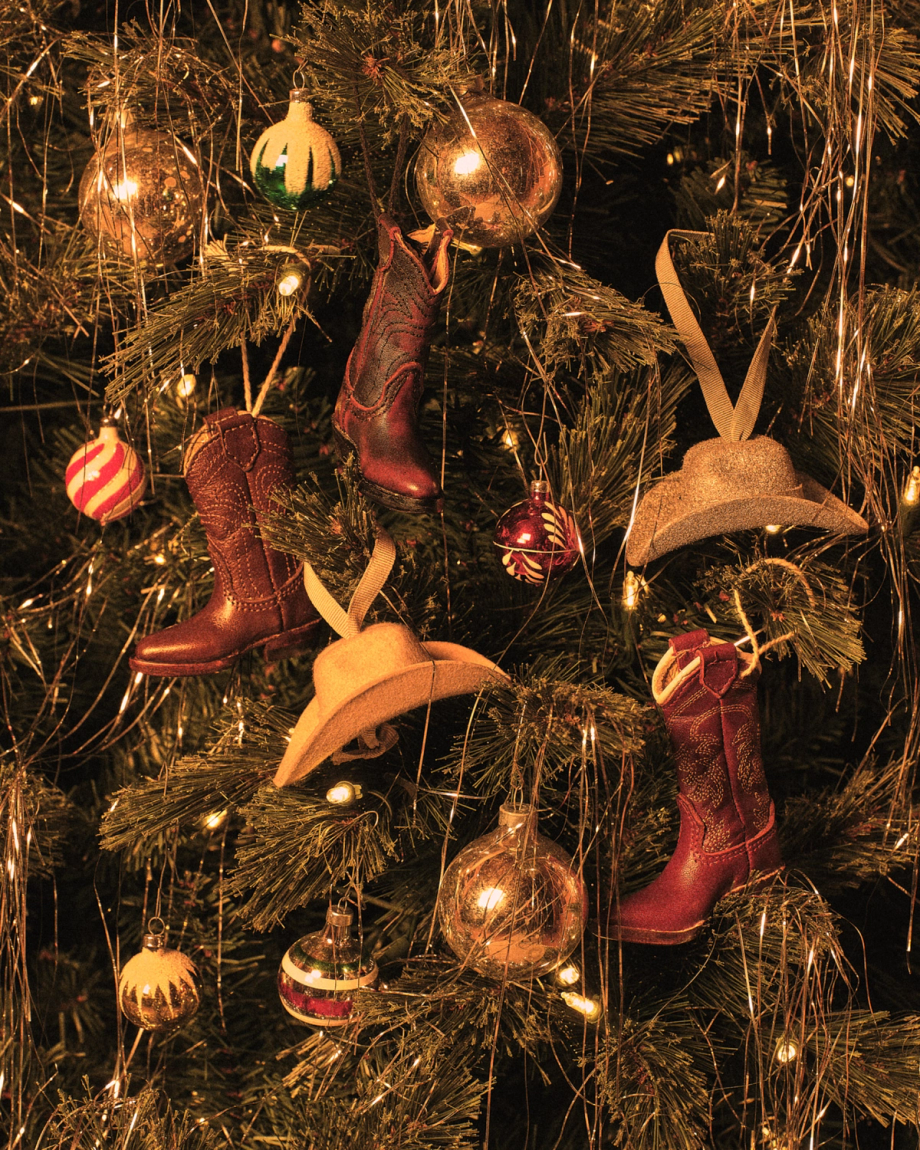 A Christmas tree decorated with cowboy boot and hat ornaments, gold and red baubles, and string lights.