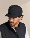 A man with a beard wears a black cap with "Tecovas Quality Made Western" text and a black vest over a white shirt, looking slightly to his left.