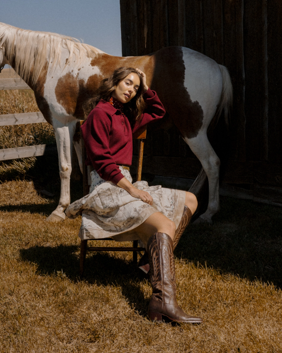 The Abby in Mocha Goat. A woman in a red sweater and skirt sits on a wooden chair next to a brown and white Mocha Goat.