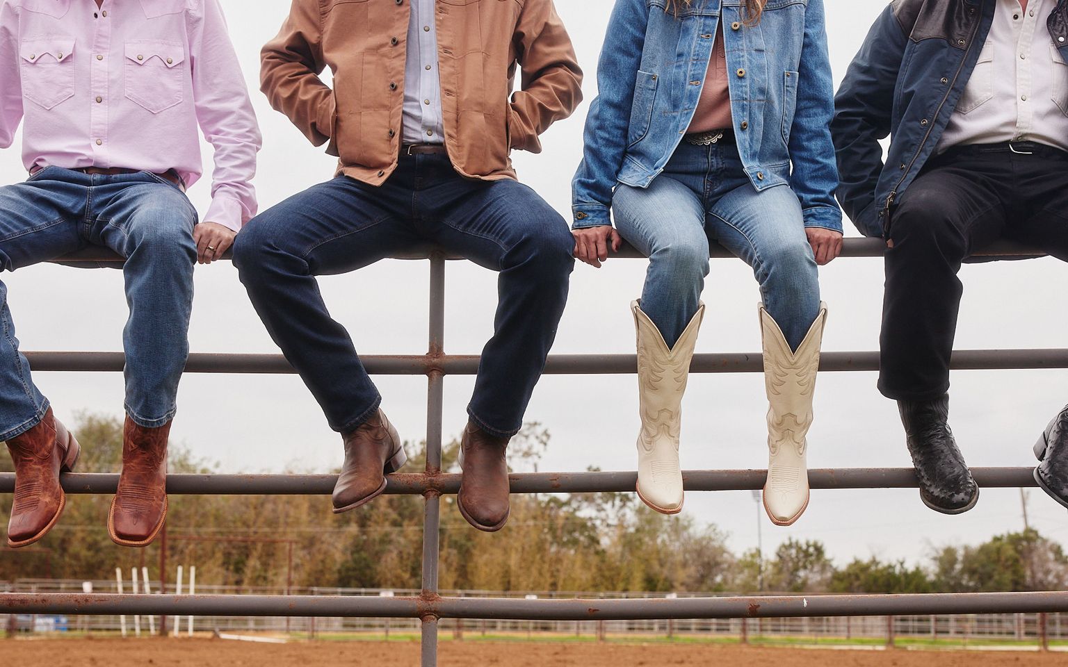 A group of people outside sitting on a rodeo fence wearing Tecovas boots and denim.