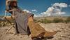 A person in a checked dress and cowboy hat sits on a wooden chair outdoors, wearing tan cowboy boots, with a desert landscape and blue sky in the background.