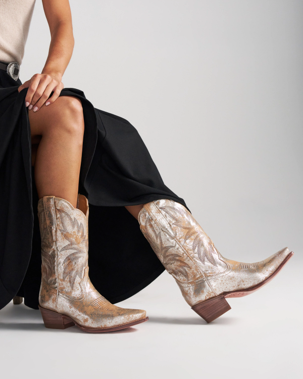 The Sadie in Platino Cowhide. Person wearing a long black skirt and metallic gold Platino western-style boots with intricate stitching.