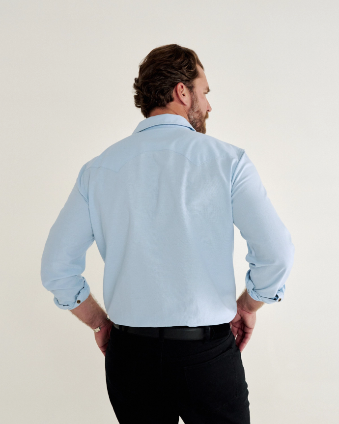 A man with short hair stands facing away, wearing a light blue button-up shirt and black pants, against a plain white background.