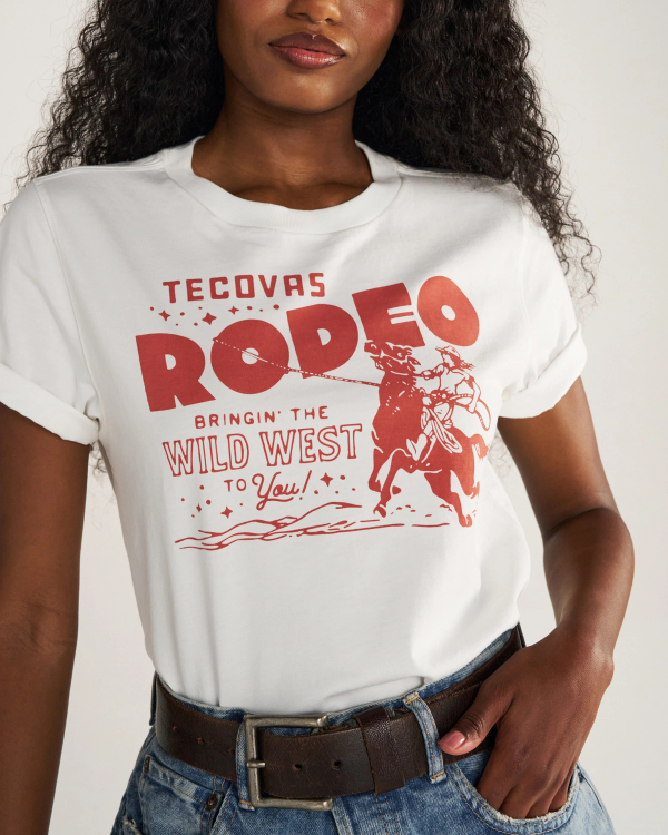A woman wearing a white T-shirt with red text and graphics that read "Tecovas Rodeo, Bringin' the Wild West to You!" She is also wearing blue jeans and a brown belt.