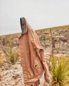 Brown trucker jacket hanging on a barbed wire fence in a desert landscape with pale blue sky and sparse vegetation.