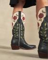 A close-up of a person wearing black cowboy boots with embroidered red roses and green leaves, standing on a beige floor.