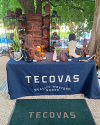 A display of cowboy boots is arranged on shelves and a table covered with a blue TECOVAS banner at an outdoor event.
