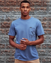 Man wearing the standard issue pocket tee in blue fin in front of a rustic wall