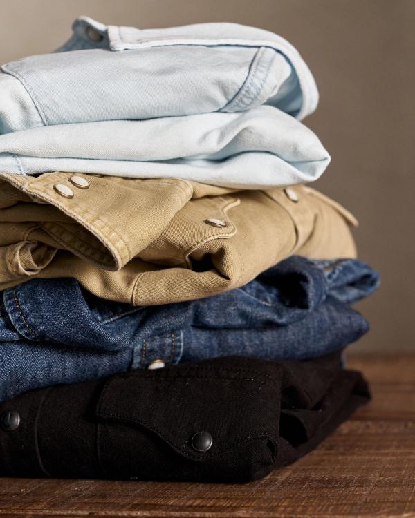 A neatly folded stack of shirts in various colors, including light blue, beige, denim blue, and black, sits on a wooden surface.