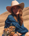 A person wearing a wide-brimmed hat and a blue dress, holding a bouquet of wildflowers with dry hillside in the background.