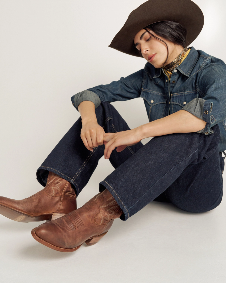 Person wearing a cowboy hat, denim shirt, jeans, and brown cowboy boots sits on the floor with eyes closed and head tilted down.